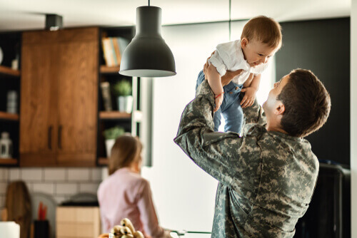 Military Servicemember holding up child with spouse in background